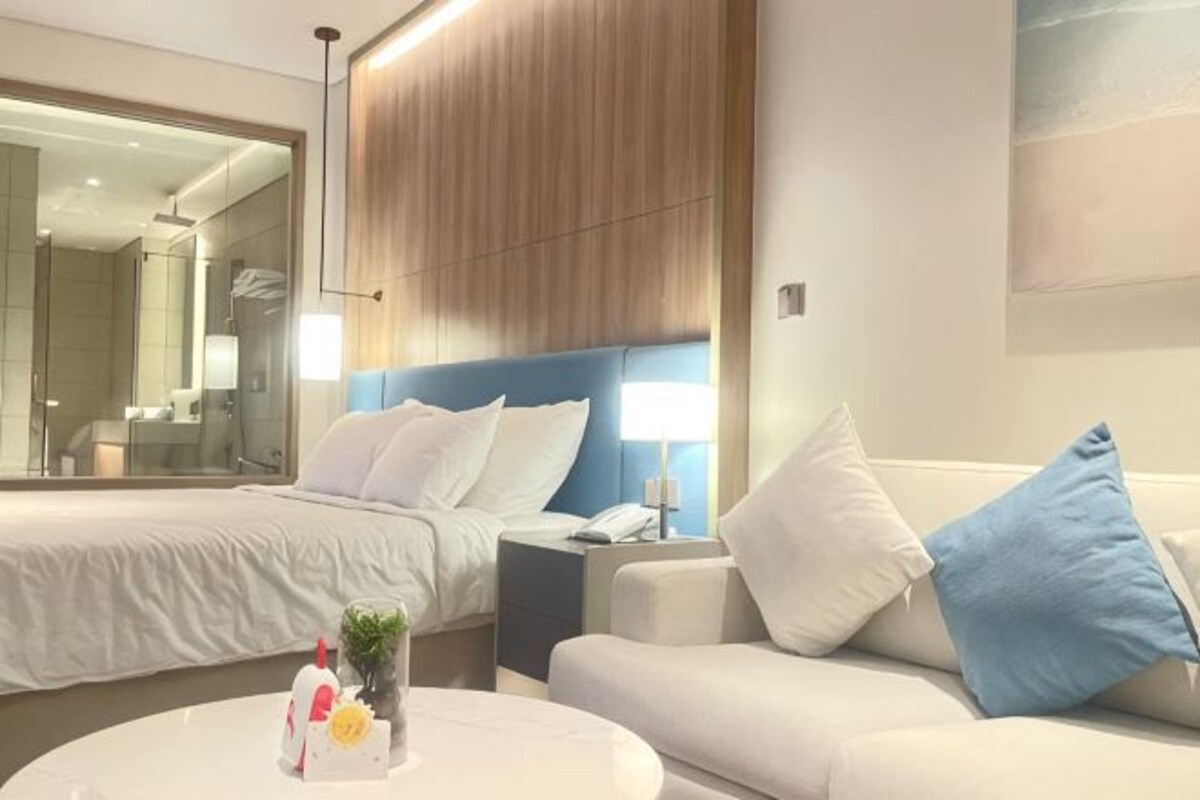 A hotel room with a bed, a sofa, a bedside table, a lamp, and a mirror.