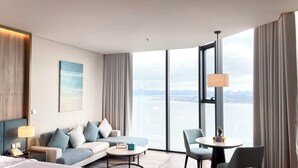 A modern hotel room with a sofa, chairs, a small table, and a large window offering a city view. - Alacarte Ha Long Bay By Azure (Bai Chay)