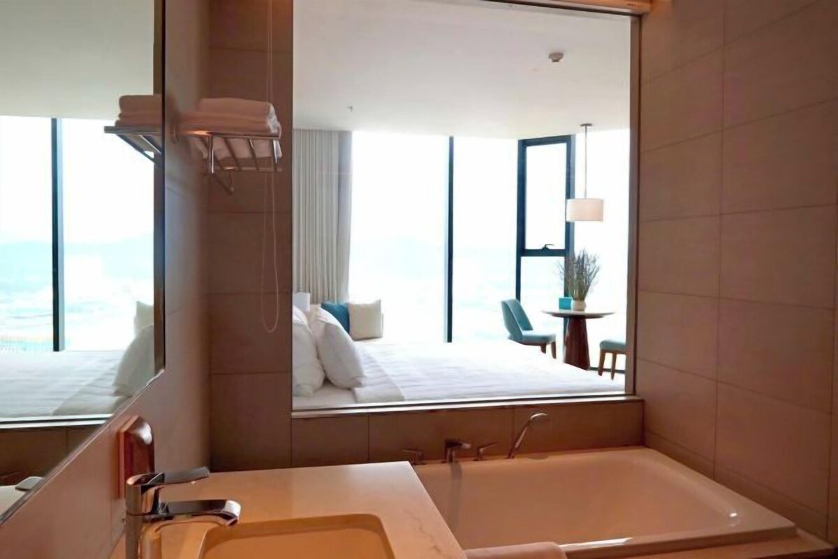 A modern hotel bathroom with a large bathtub, a mirror, and a view of a bedroom with a bed and a table.