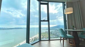 A room with a large window offering a view of a beach and cityscape. - Alacarte Ha Long Bay By Azure (Bai Chay)