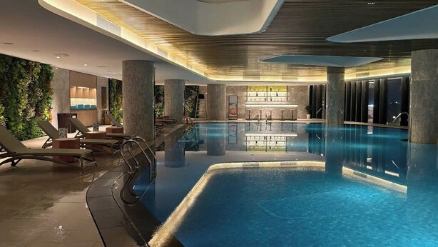 A modern indoor swimming pool with clear water, surrounded by lounge chairs and a reception area with a bar. - Alacarte Ha Long Bay By Azure (Bai Chay)