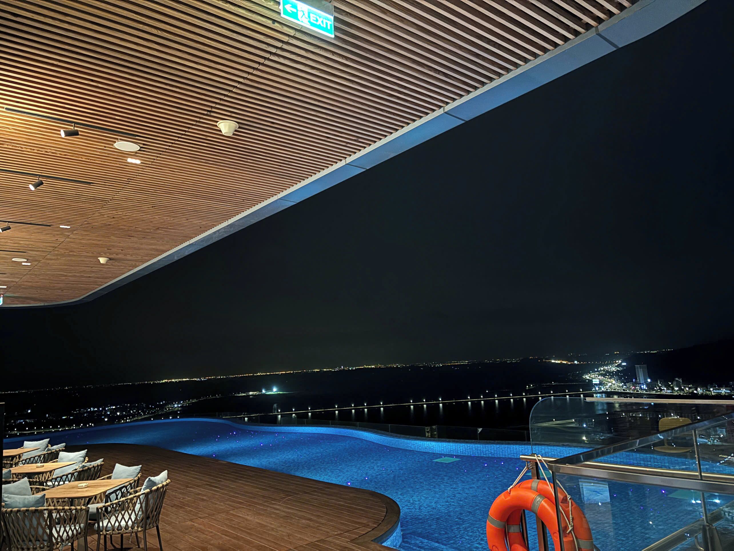 A pool area with a wooden ceiling, a lifebuoy, and a view of a cityscape at night.