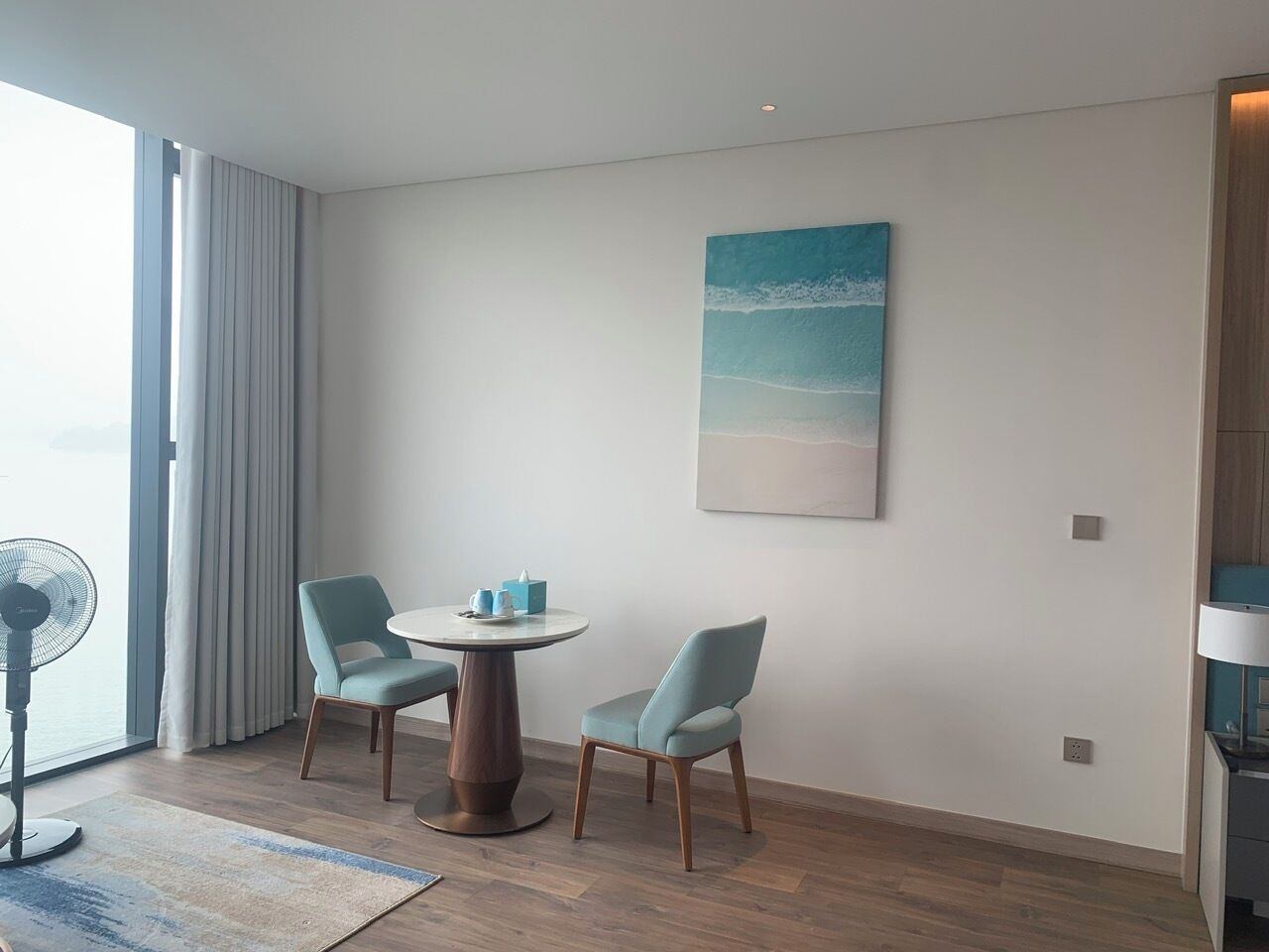 Une chambre d’hôtel moderne avec une table ronde et deux chaises, une grande fenêtre avec des rideaux, un ventilateur et un tableau représentant une scène de plage.
