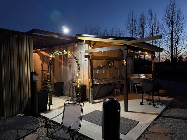 An outdoor patio area with a wooden pergola, a metal chair, a table, and a trash can.
