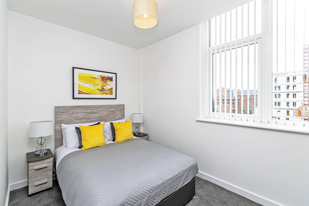 A modern hotel room with a large bed, a nightstand, a lamp, a framed picture, and a window with blinds.