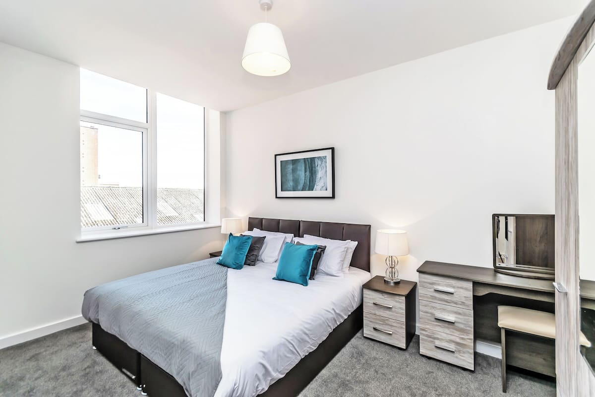 A modern bedroom with a large bed, a desk, and a window with a view of buildings.