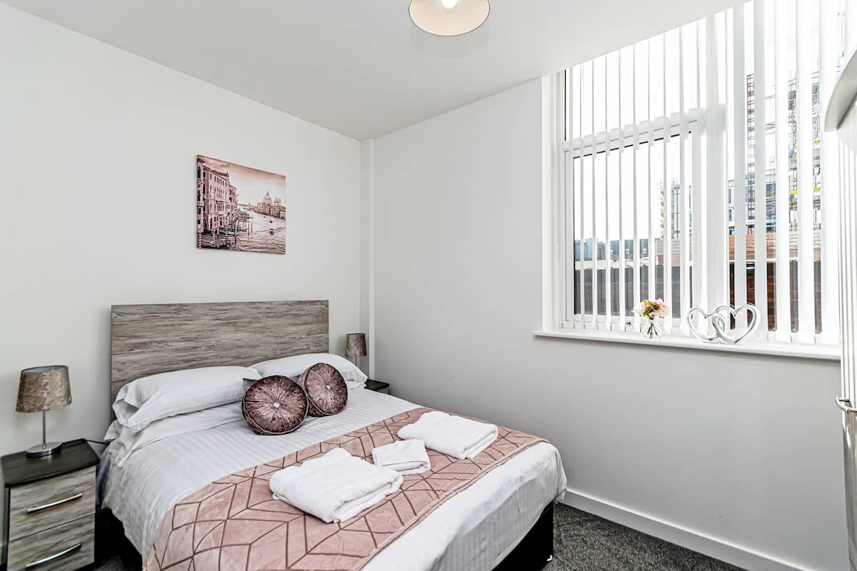 A neatly arranged bedroom with a bed, bedside tables, a window with blinds, and a cityscape painting on the wall.