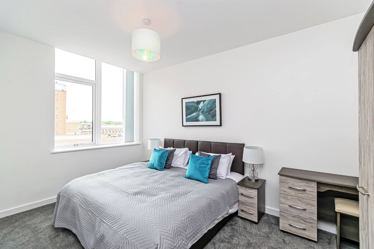 A bedroom with a large bed, a desk, a chair, a nightstand, and a window with a view of buildings.