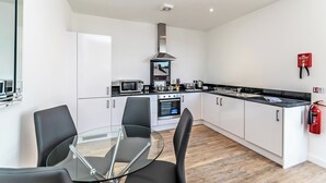 A modern kitchen with white cabinets, a stainless steel range hood, a microwave, and a built-in oven. There is a glass table with chairs, a fire extinguisher on the wall, and a fire blanket mounted on the wall. - City Suites by Guestz (Chester)