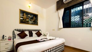 A hotel room with a bed, a nightstand, a wall-mounted air conditioner, a window with curtains, and a framed picture on the wall.