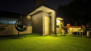A small, well-lit outdoor area with artificial grass, a hanging chair, a small table with chairs, and a door.