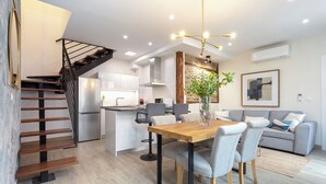 Eine moderne Küche mit einer zentralen Kochinsel, einem Essbereich mit Holztisch und Stühlen, einem grauen Sofa und einer Treppe mit schwarzer Geländer.