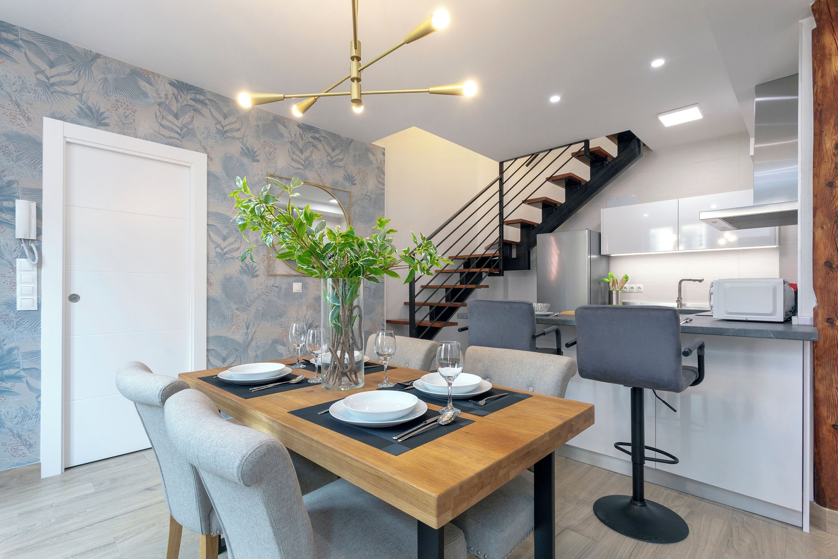 A modern kitchen with a wooden dining table set for two, a bar stool, and a staircase leading to an upper level.