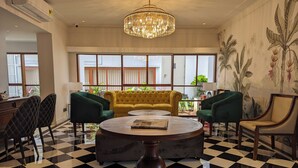 A hotel lobby with a checkered floor, a yellow tufted sofa, green armchairs, a round coffee table, and a chandelier.