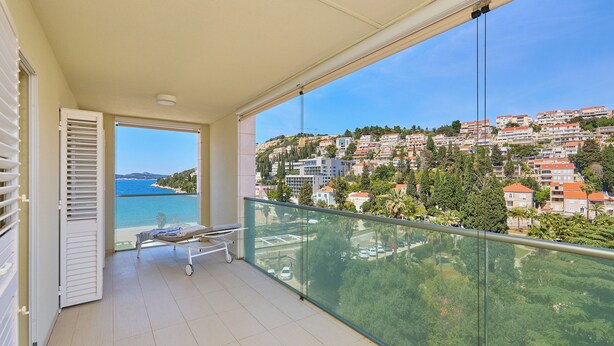 A balcony with a view of a coastal town, a glass railing, and a small table with a chair.