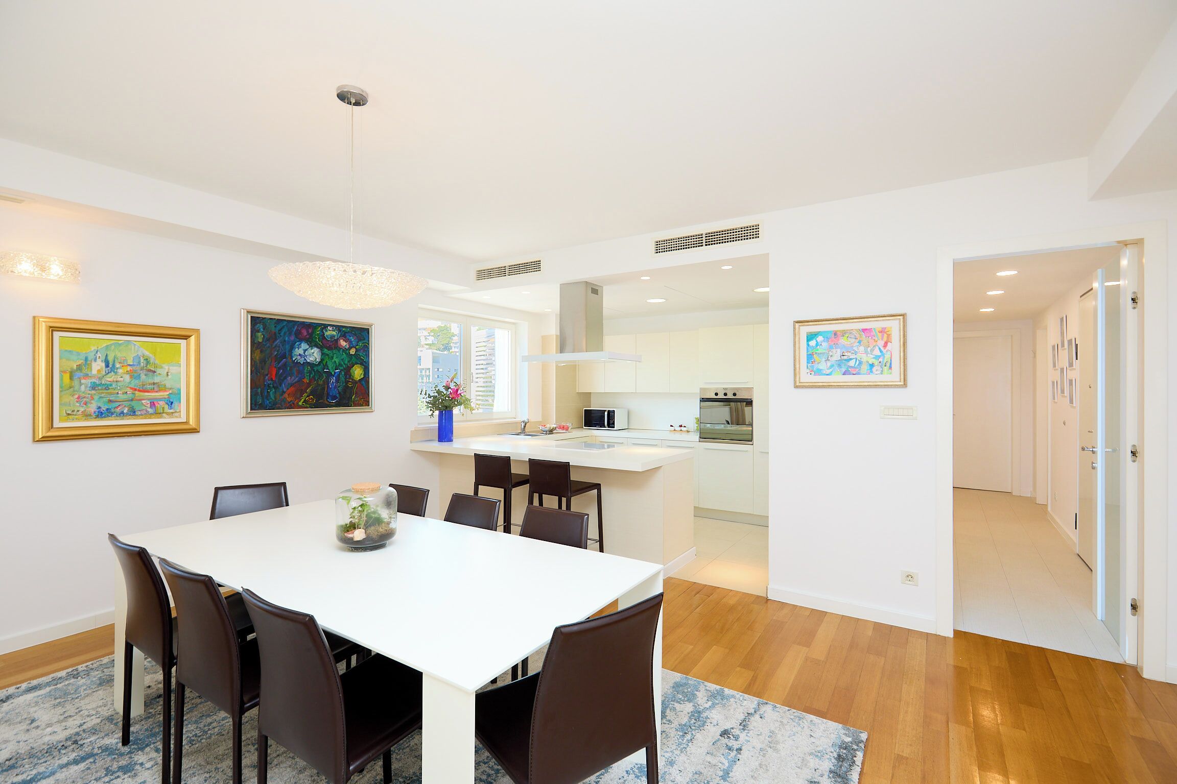 A modern dining area with a white table and brown chairs, a kitchen with white cabinets, and artwork on the walls.