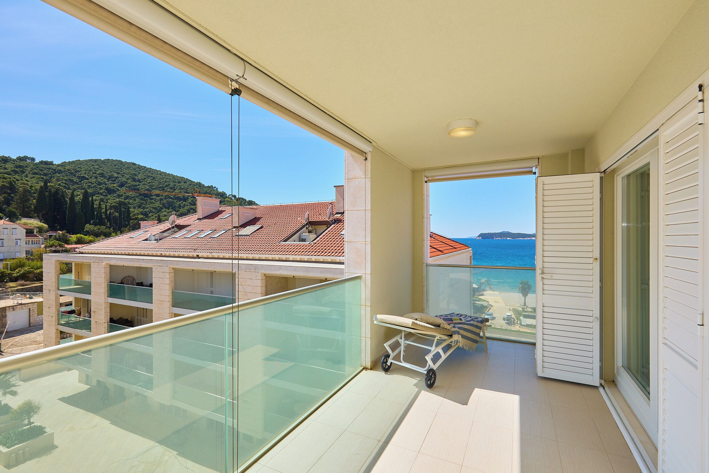 Ein Balkon mit Glasgeländer, Blick auf den Strand und ein Liegestuhl.