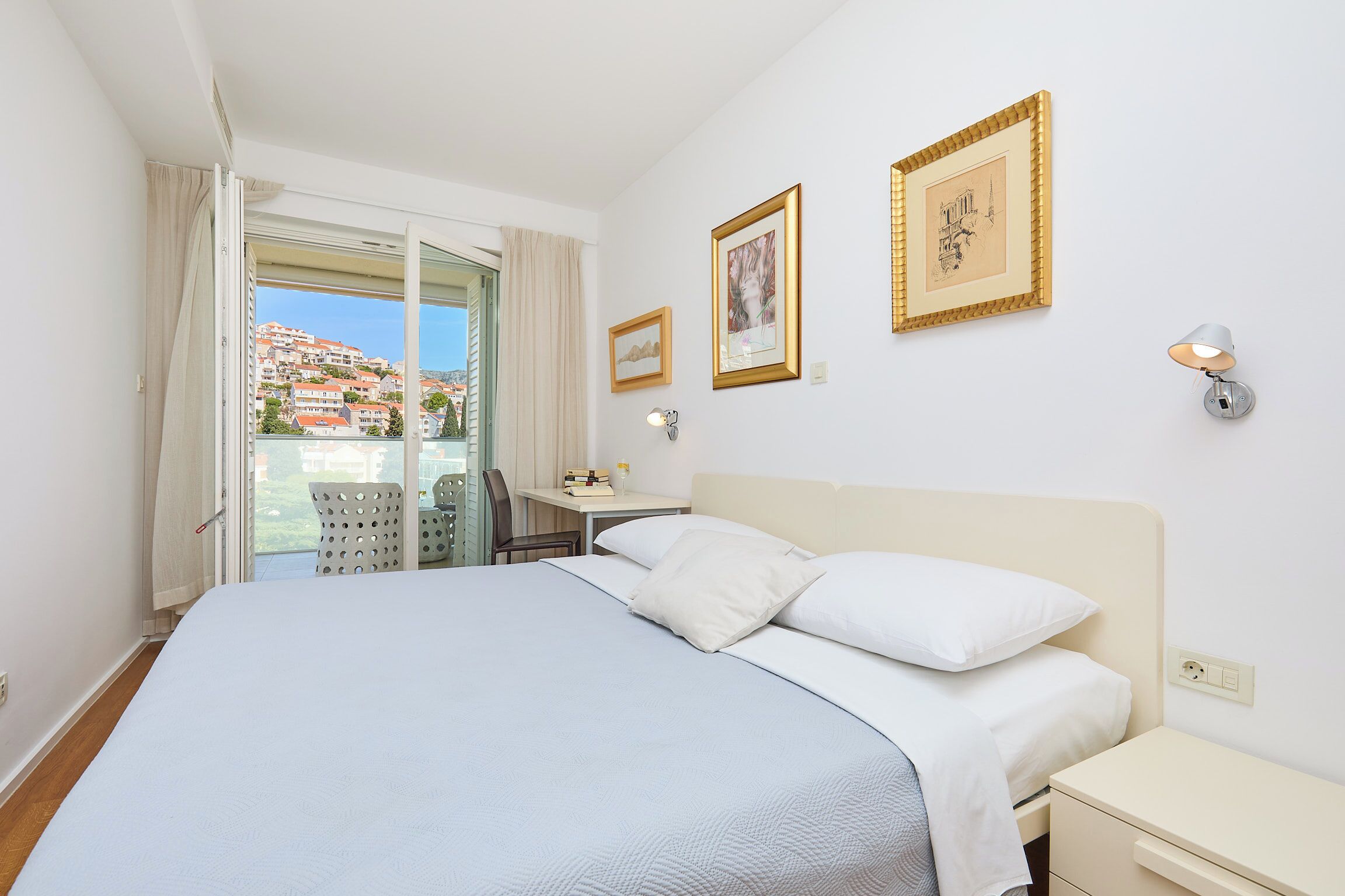 A hotel room with a bed, a desk, a chair, and a balcony with a view of buildings.