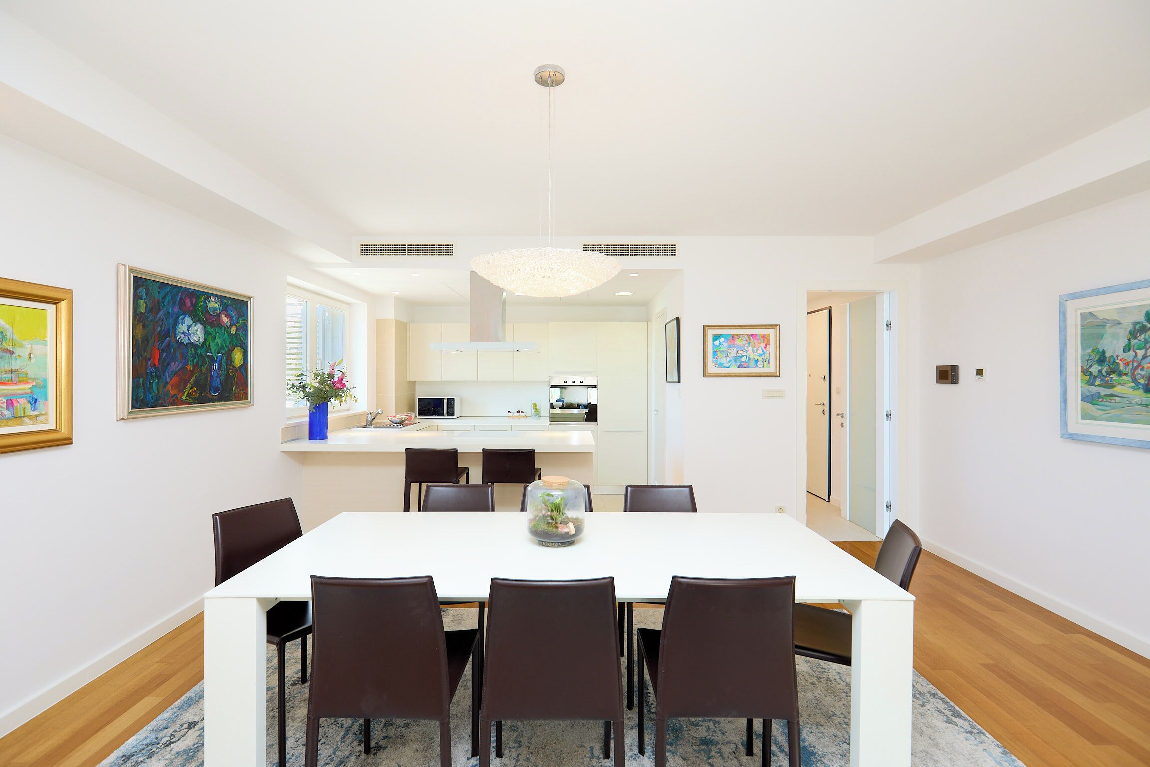 A modern dining area with a white table and dark chairs, a kitchen in the background, and artwork on the walls.