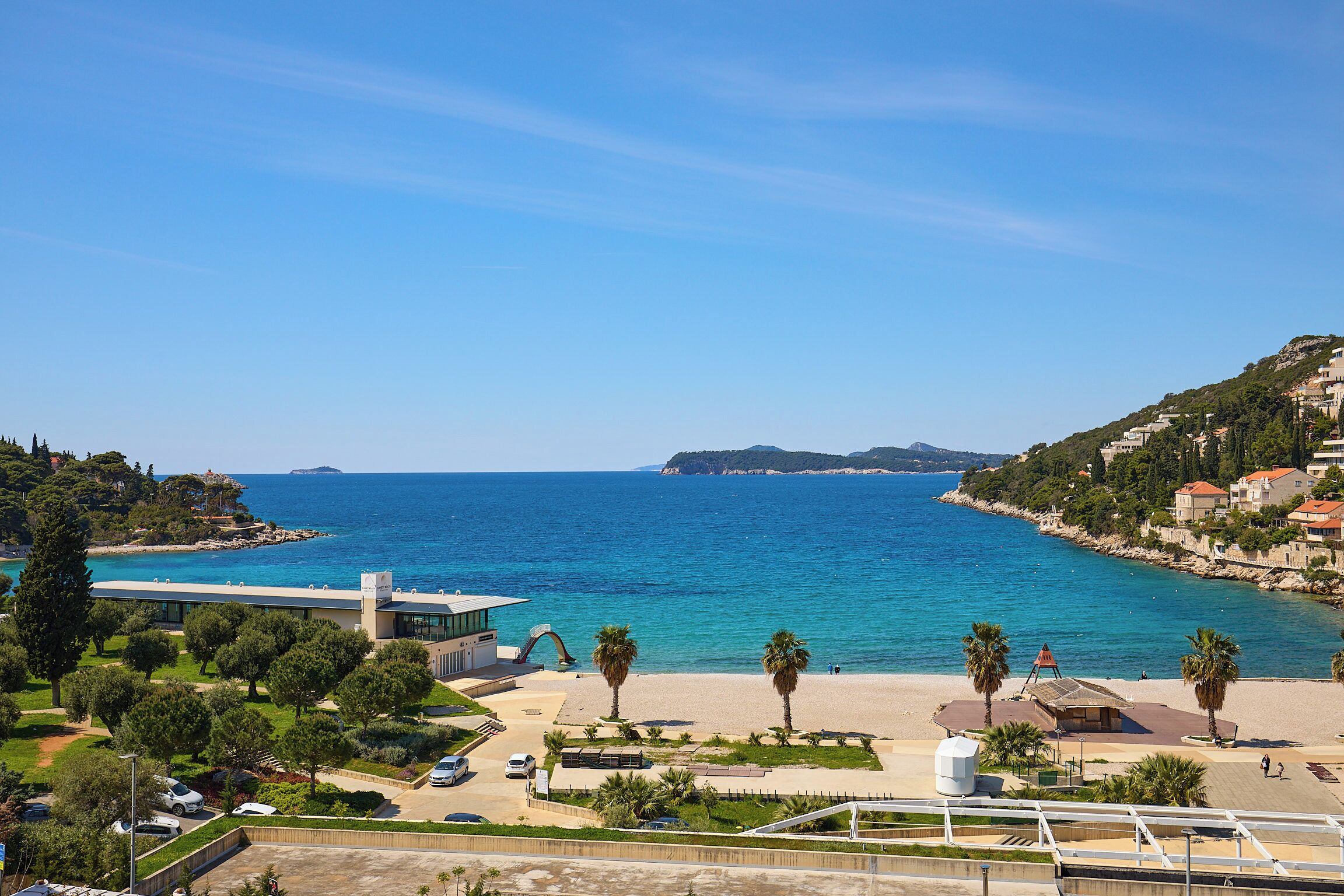 A coastal view with a beach, palm trees, and a modern building with a glass facade.