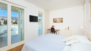 A hotel room with a bed, a desk, a chair, a TV, and a window with a view of buildings and a mountain.