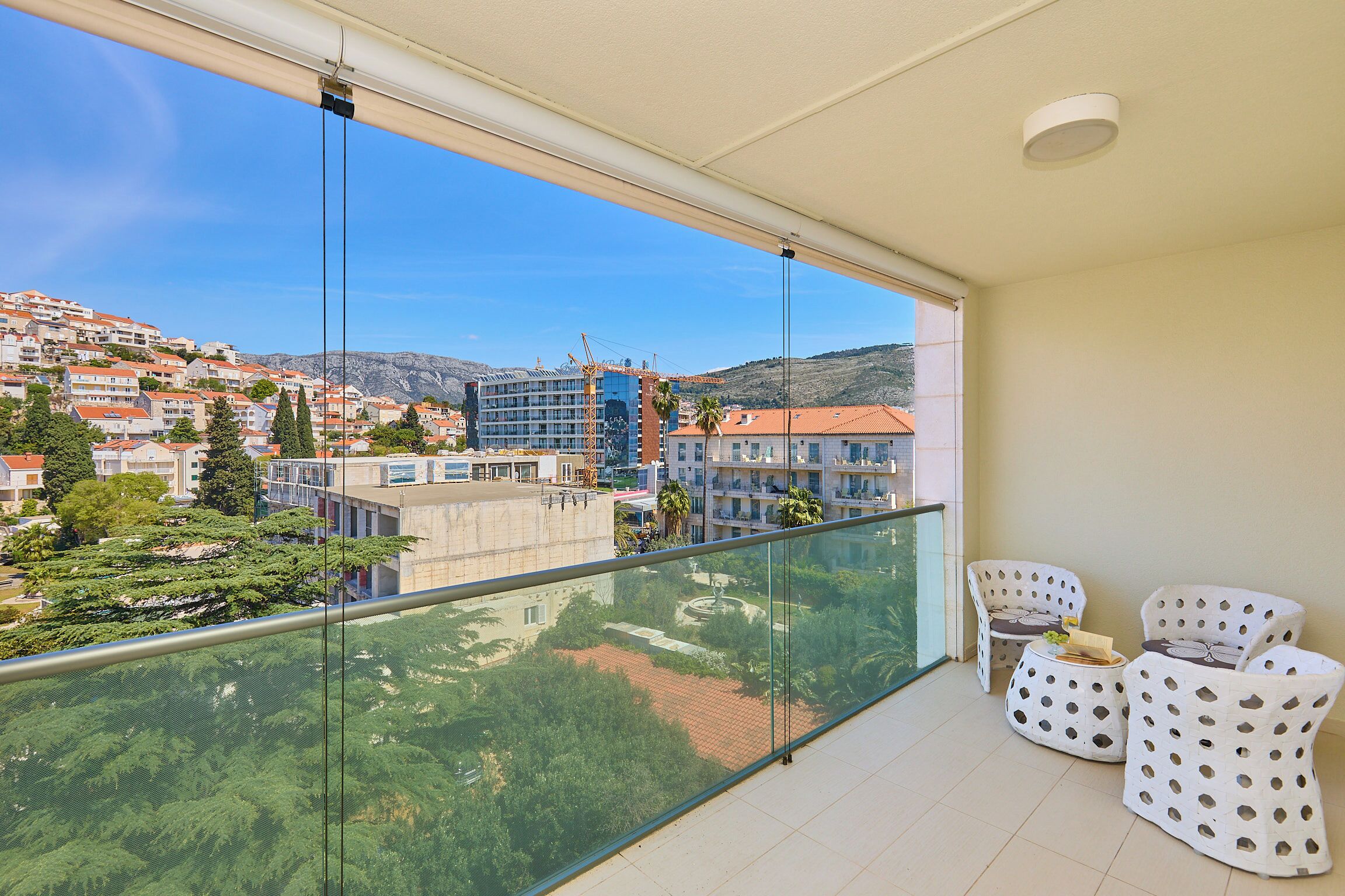 Un balcon doté d’une balustrade en verre, de chaises blanches à pois et offrant une vue sur une ville perchée sur une colline, avec ses bâtiments et ses arbres.