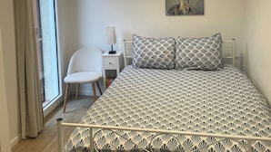 A bedroom with a bed, a chair, a nightstand, a lamp, a painting, and a window with curtains.