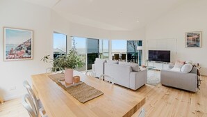 Uma sala de estar espaçosa com uma mesa de jantar de madeira, dois sofás e uma televisão. Grandes janelas proporcionam vista para o mar.
