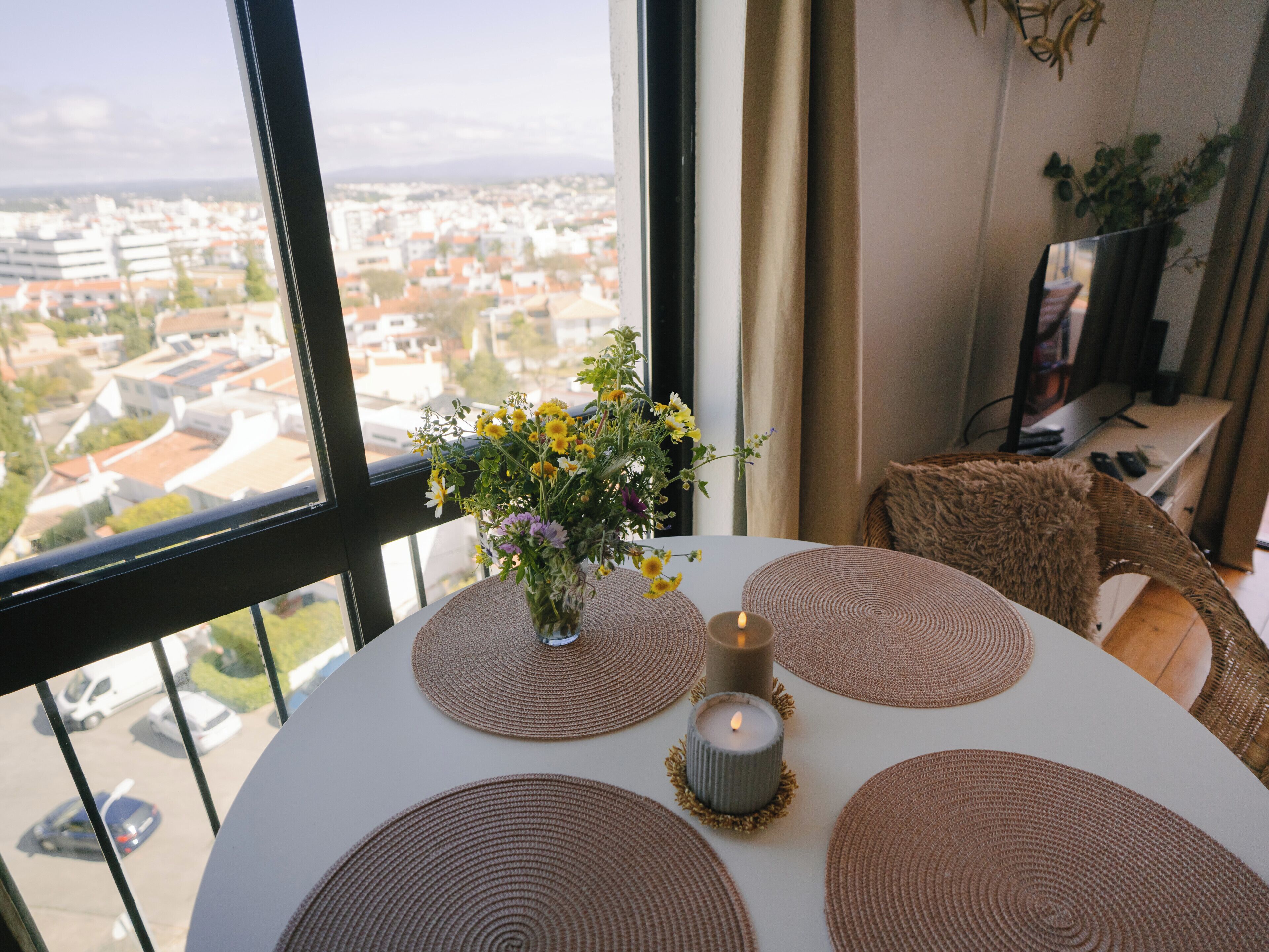 A room with a large window overlooking a cityscape, a table with placemats, a lit candle, and a vase with flowers.
