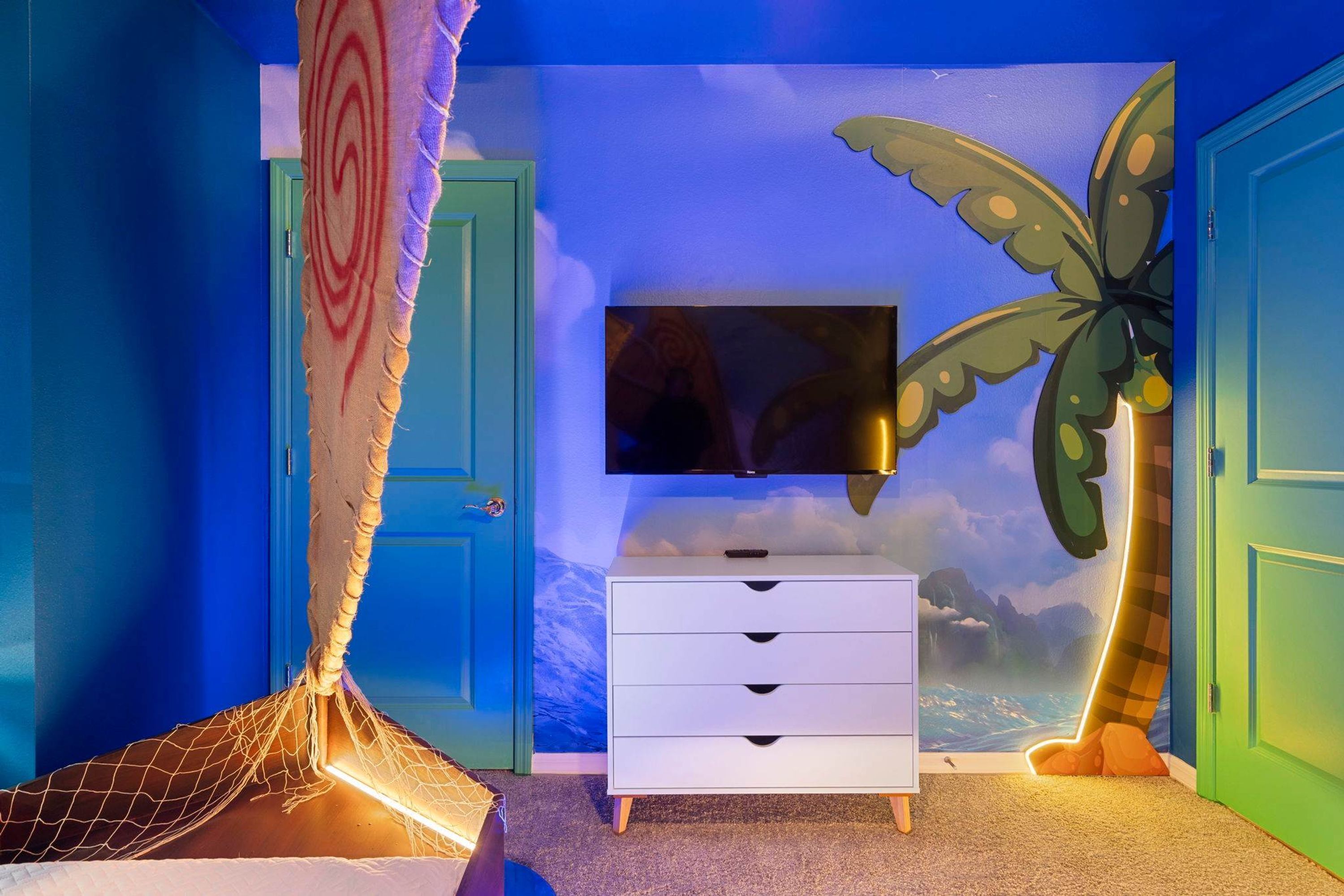 Une chambre à coucher avec un lit, une commode blanche, un téléviseur et une fresque représentant un paysage tropical.