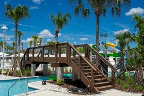 A pool area with a wooden bridge, palm trees, and a water slide. - 1149 Spacious Family 5-bedroom Townhome (Clermont)