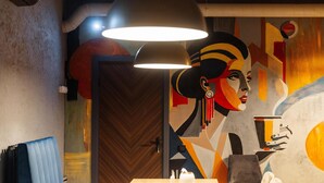 A room with a wooden herringbone floor, a table with a white kettle, and a chair. There is a door with a wooden panel and a wall with a colorful abstract mural. - Avenue Hotel (Uralsk)