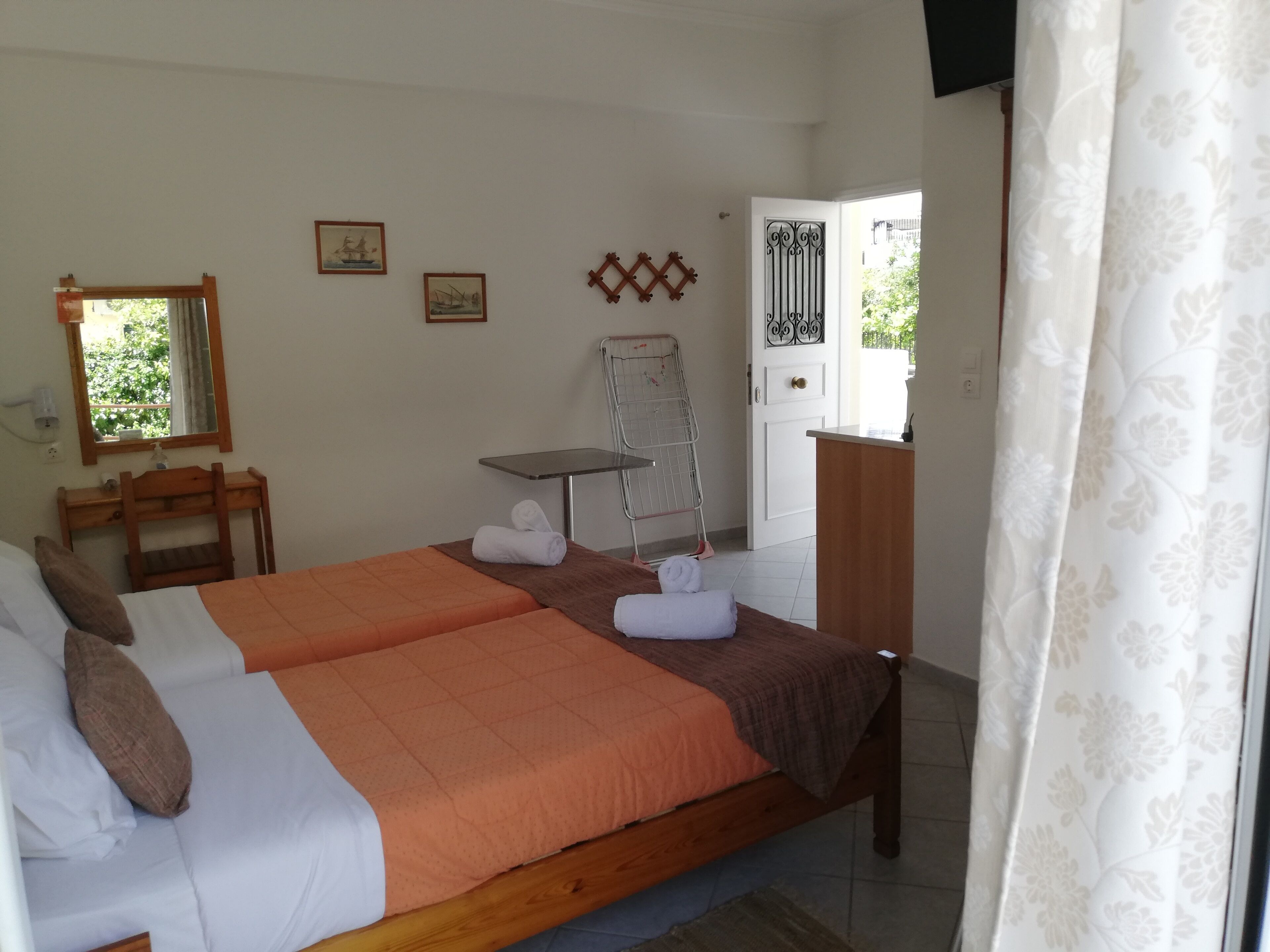 A double bed with orange bedding, a wooden desk, a chair, a small table, a drying rack, and a door with a decorative iron gate.