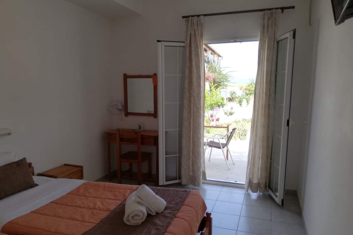 A bedroom with a bed, a desk, a chair, and a view through a door to an outdoor area.