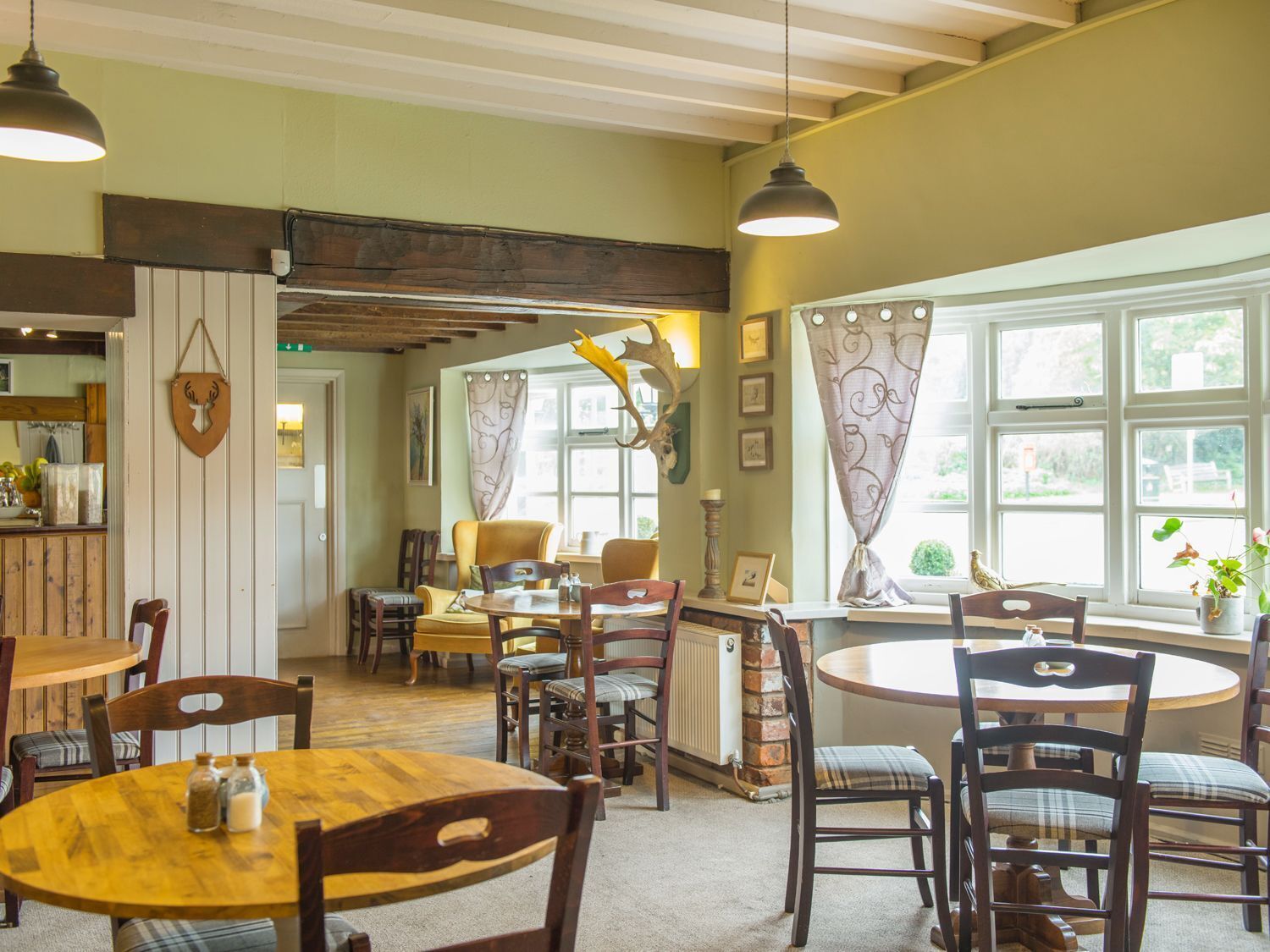 A cozy dining area with wooden tables and chairs, a rustic ceiling, and large windows with curtains.