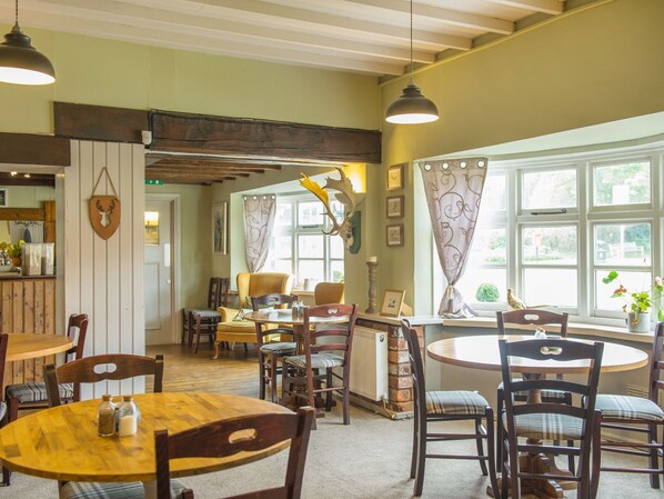 Un coin repas chaleureux avec des tables et des chaises en bois, un plafond rustique et de grandes fenêtres ornées de rideaux.