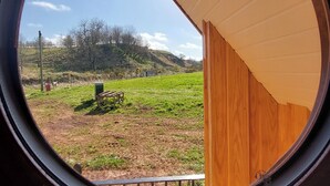 Blick durch ein rundes Fenster in einen Raum mit Holzbalkendecke und eine begrünte Außenanlage mit Bank und Bäumen.
