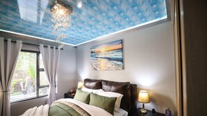 A bedroom with a bed, nightstands, a lamp, a painting, and a view of the outdoors through a window.