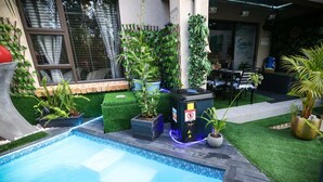 A swimming pool with surrounding greenery, a seating area, and a building with a window.