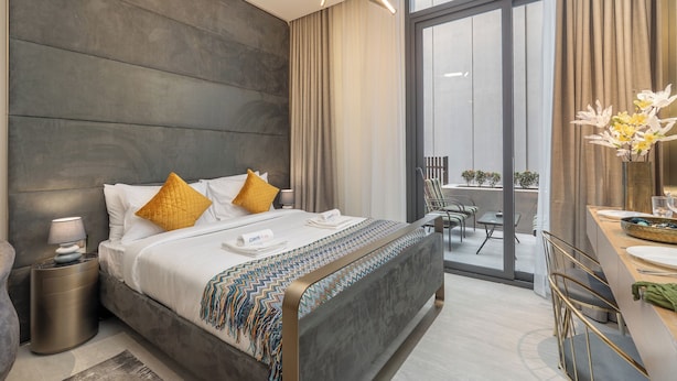 A modern hotel room with a large bed, a bedside table, a desk, and a balcony with outdoor seating. - Czechin - Modern Studio JVC  (Dubai)