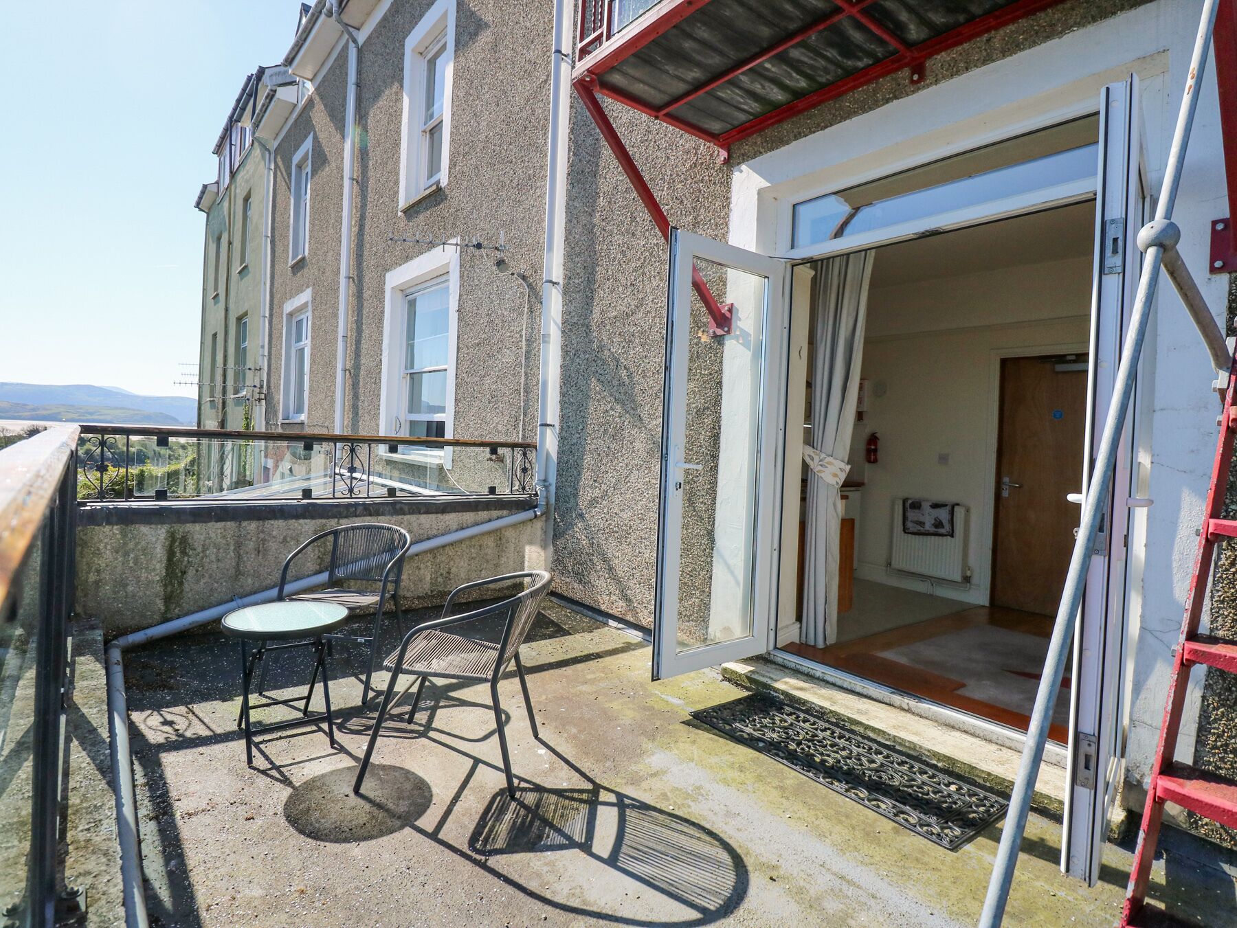A balcony with two chairs and a table, a red ladder, and a door leading inside.