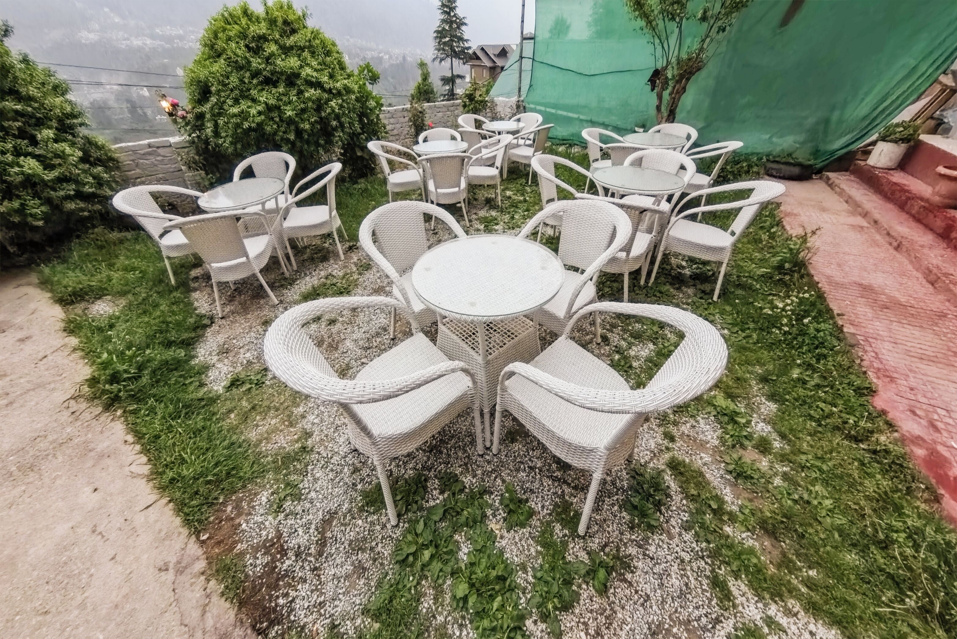 Un patio con mesas redondas y sillas de mimbre blanco, rodeado de follaje y un muro de piedra.