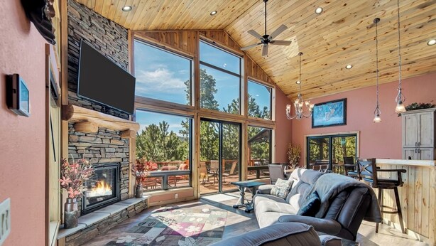 A spacious living room with a stone fireplace, large windows, a ceiling fan, and a cozy seating area. - Wolf Ridge- Deadwood South Dakota (Sturgis)