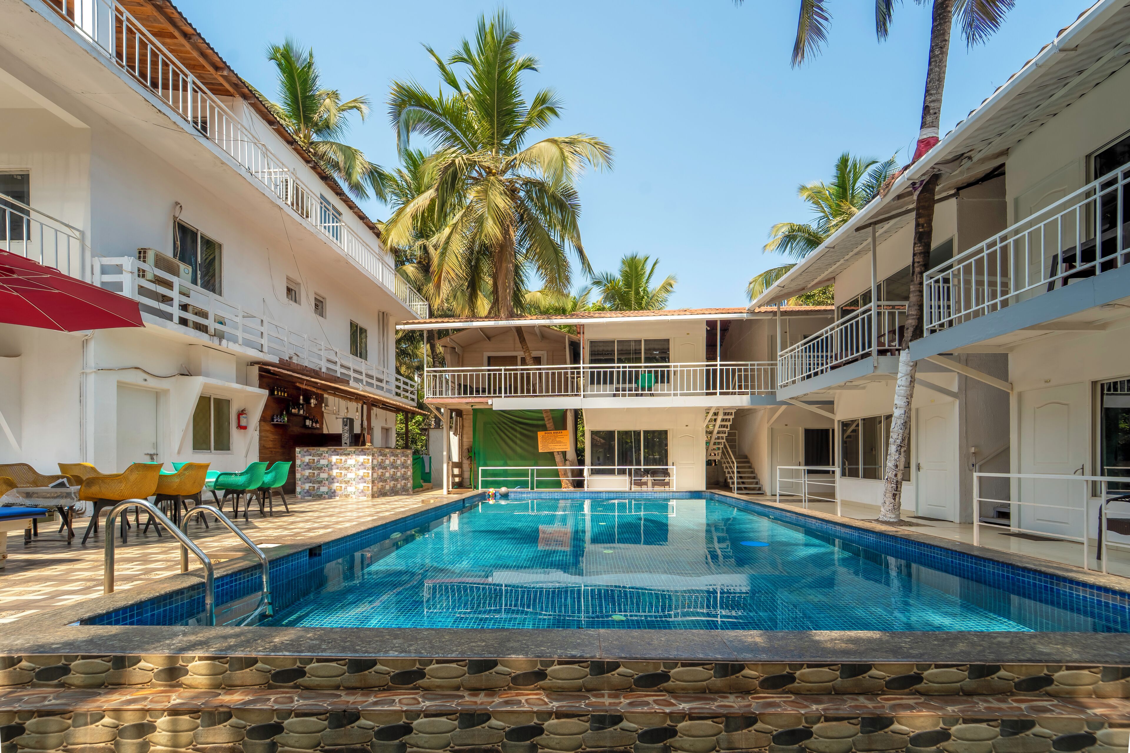 A swimming pool with lounge chairs and a bar area, surrounded by buildings with balconies and palm trees.