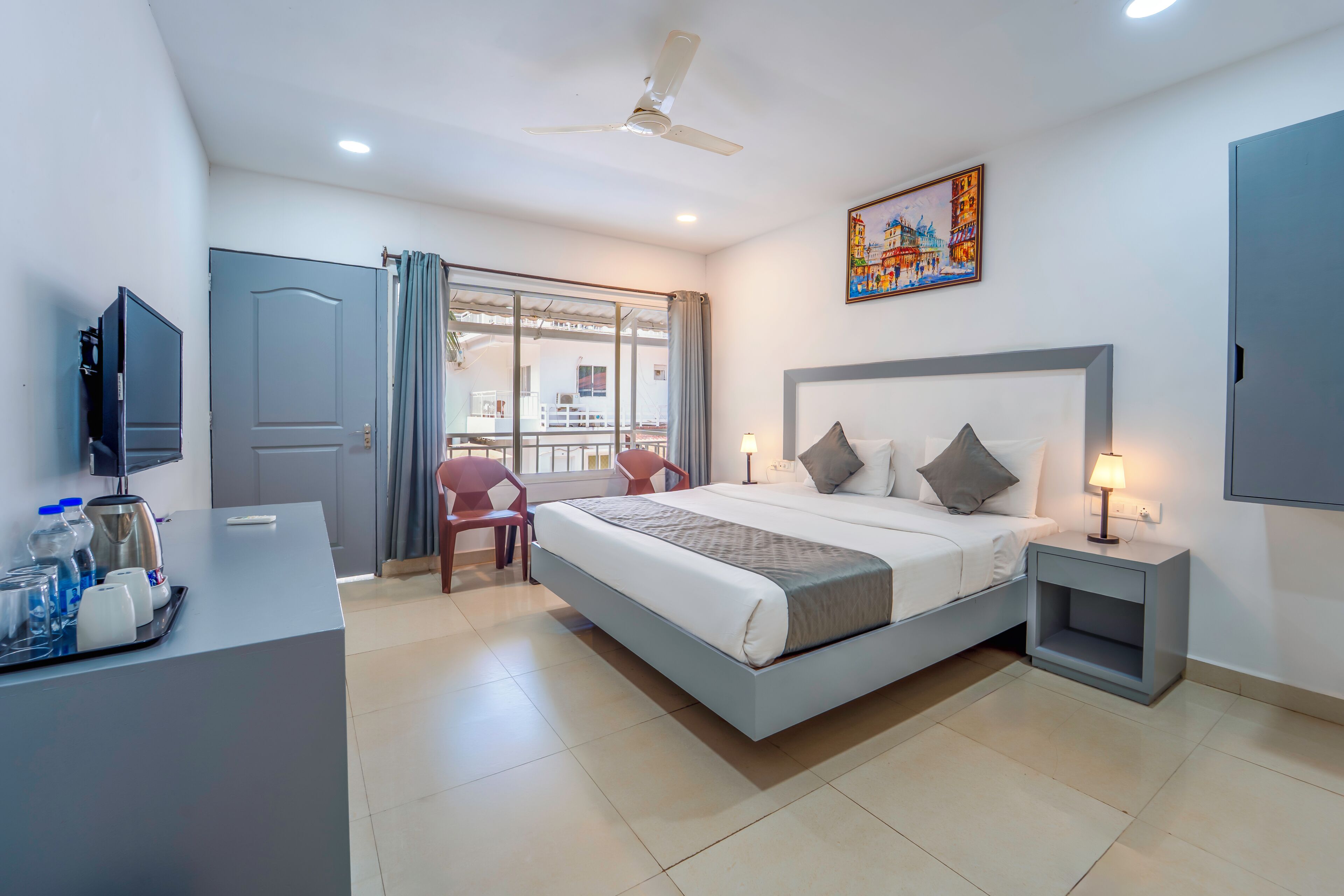 A hotel room with a bed, bedside table, TV, and a desk with a coffee maker and water bottles.