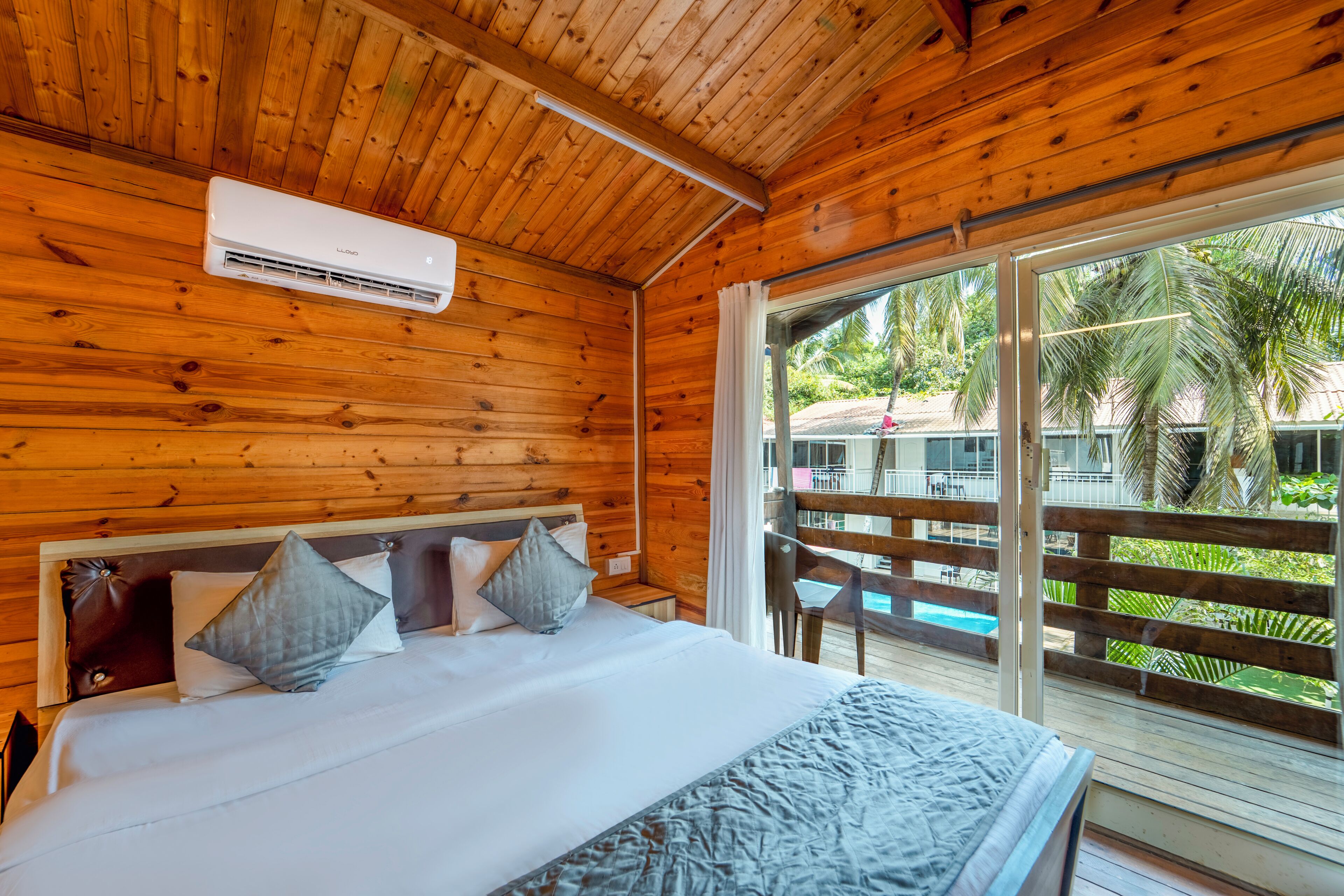 Een houten kamer met een bed, een airconditioner en uitzicht op een zwembad en palmbomen.