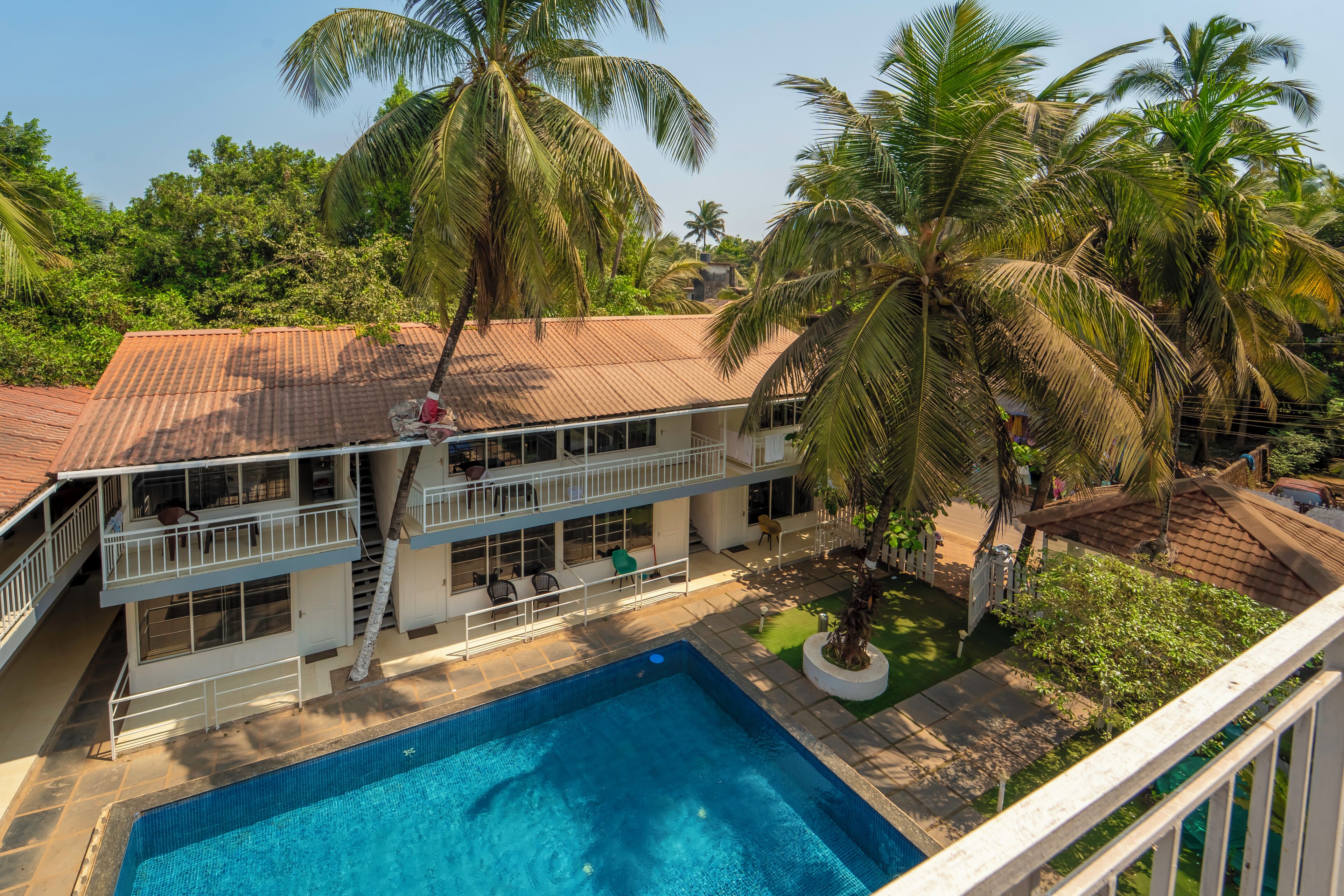 A two-story building with a swimming pool, surrounded by palm trees and greenery.