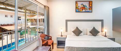 A hotel room with a bed, a desk, a chair, a nightstand, a lamp, and a large window with a view of a pool and buildings.
