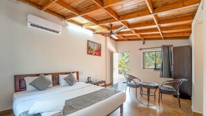 A hotel room with a bed, a ceiling fan, a painting, a window, and a balcony with chairs. - BeachWalk By FeatherTouch Hotels Resorts (Morjim)