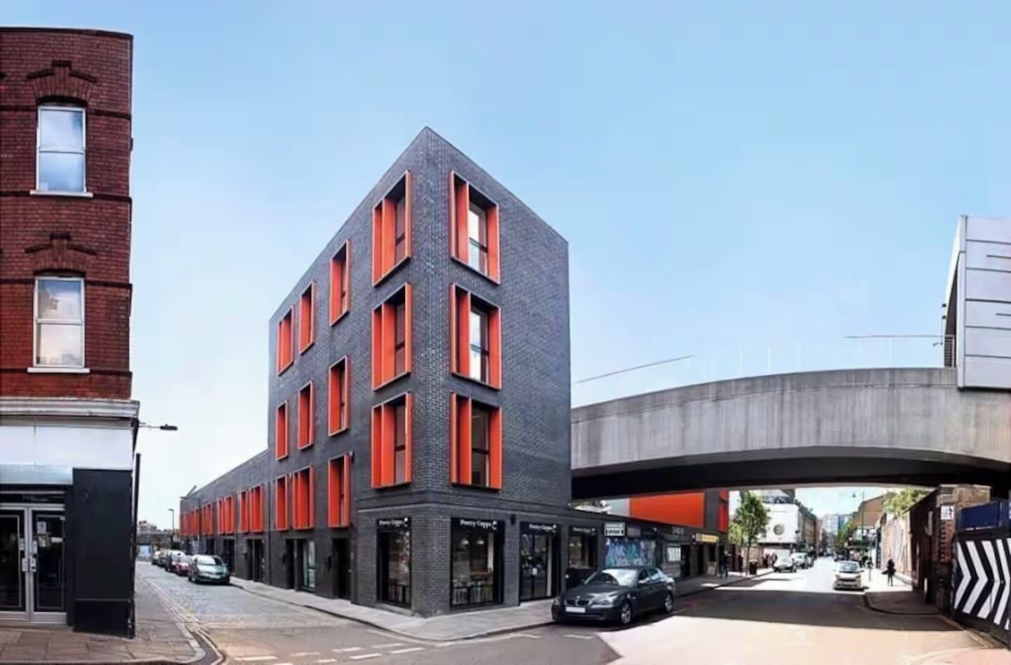 A modern building with a brick facade and red-framed windows, located on a street with a pedestrian walkway and an overpass in the background.